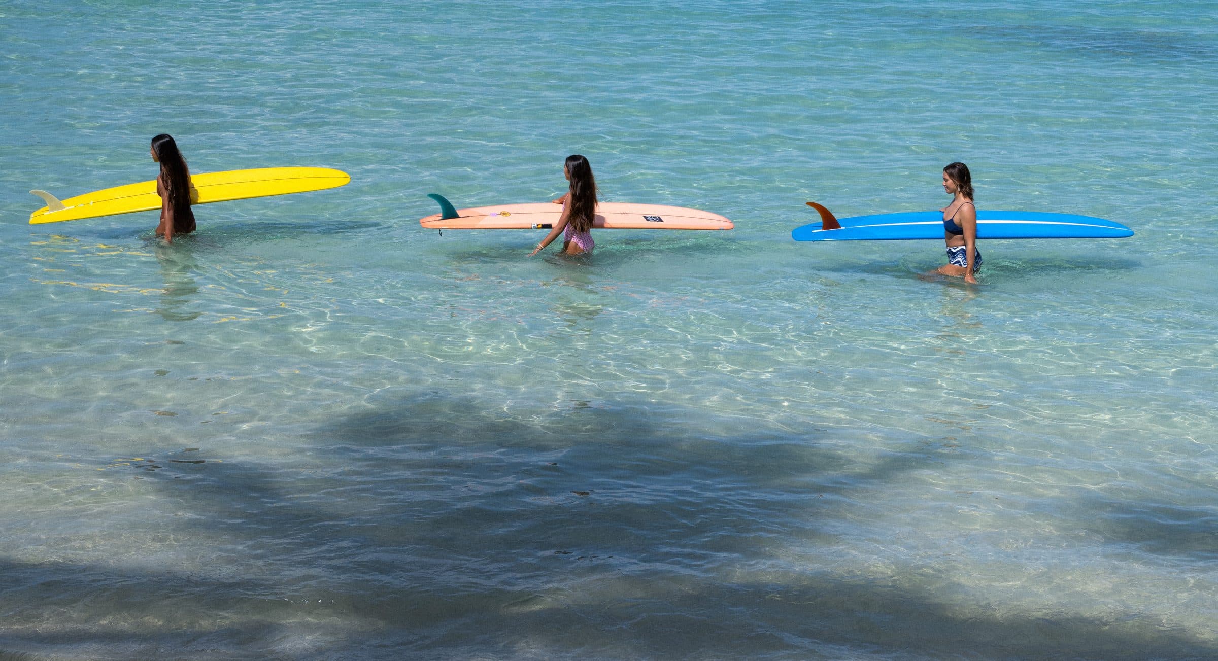Three surfers carrying longboards through clear turquoise water at Waikīkī, Oʻahu — Bluhour campaign by Develope'